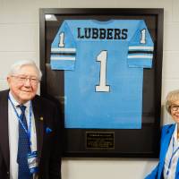 Don and Nancy Lubbers posing in front of a framed Lubbers jersey in the Jamie Hosford Football Center.
